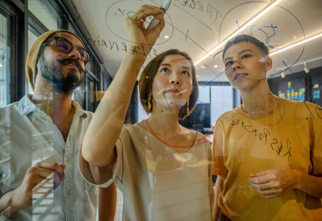 Three people stand at a glass panel and draw on it using whiteboard markers.