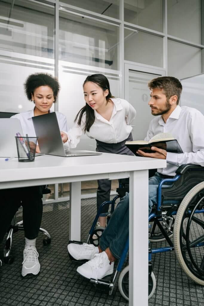 Three people gather around a laptop on a table: one sitting, one standing, and one in a wheelchair.