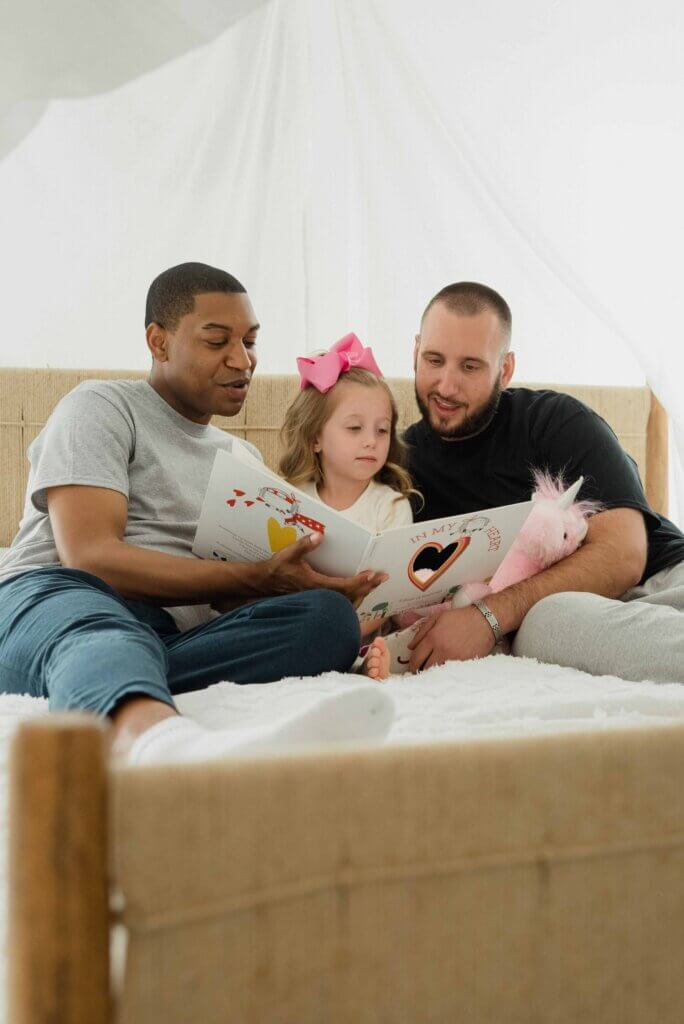 Two men sit on a couch and read a children's book to a young girl.