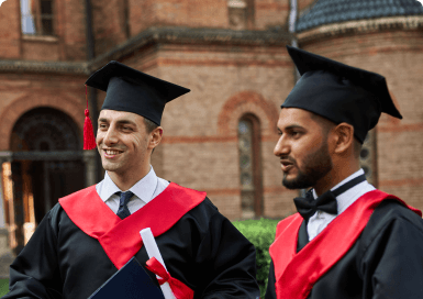 Two young men wearing graduation robes and hats stand in front of an old building